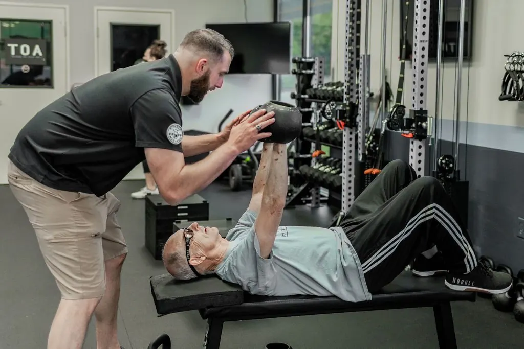 A STRONG personal trainer guiding a member through a kettlebell press